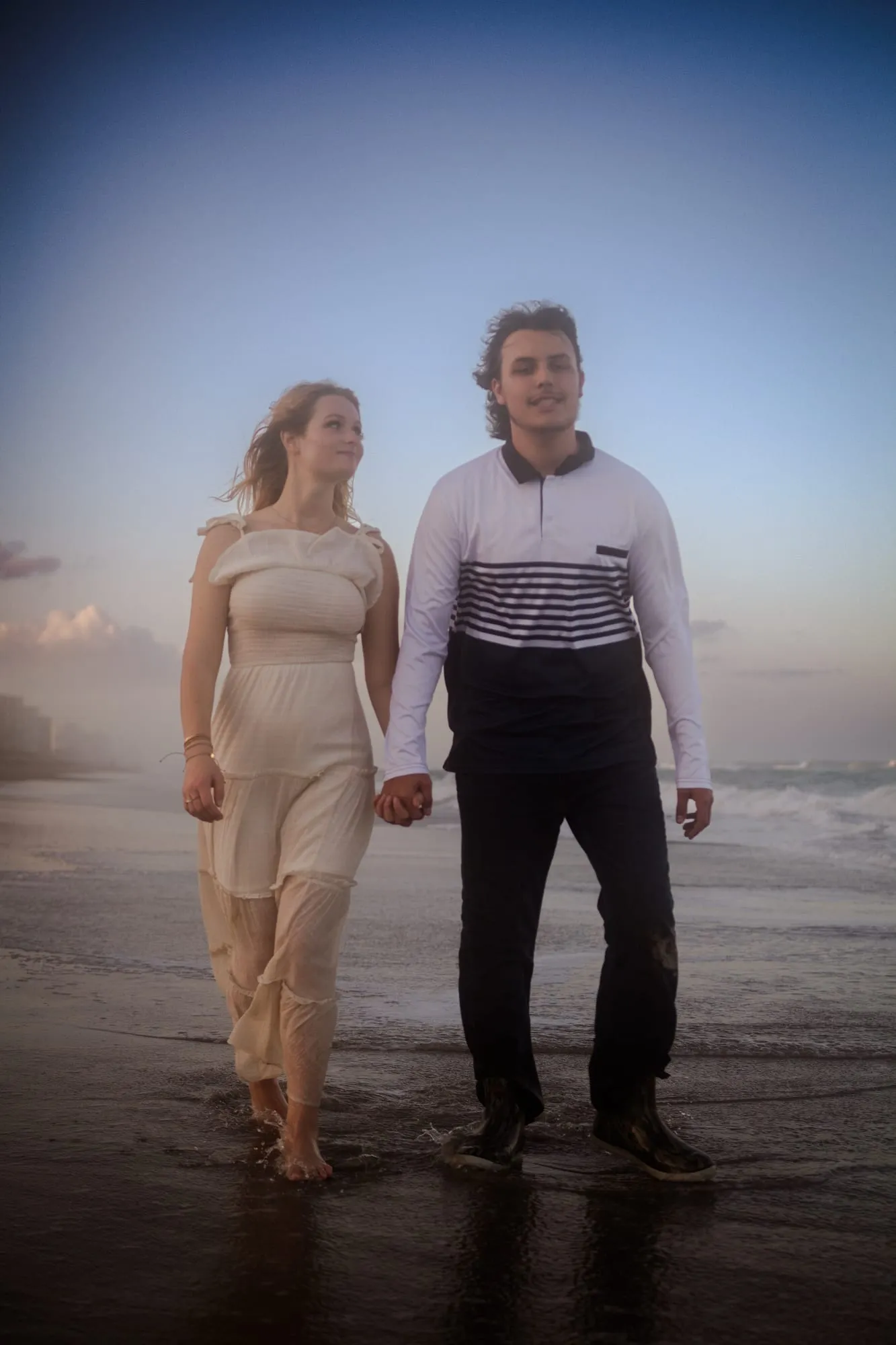 A couple walking hand in hand along the shoreline at dusk with soft sea mist rolling in behind them