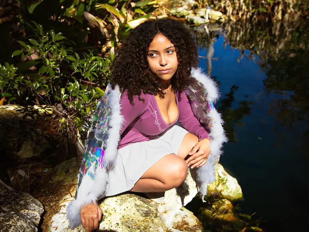 A young woman in iridescent feathered angel wings crouching on a rock beside a clear Florida spring