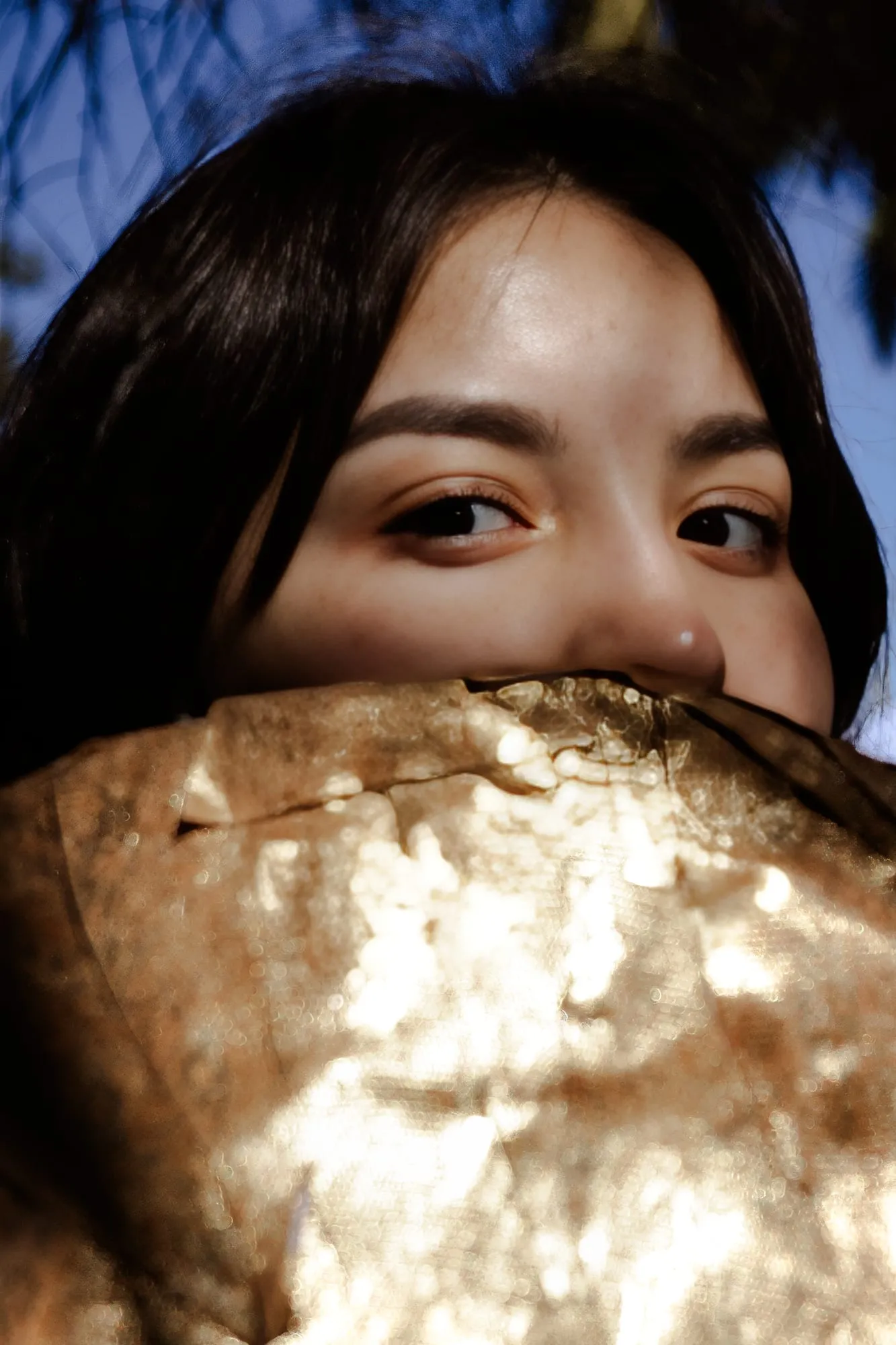 Close-up editorial portrait of a young woman framed by shimmering gold fabric in soft low light