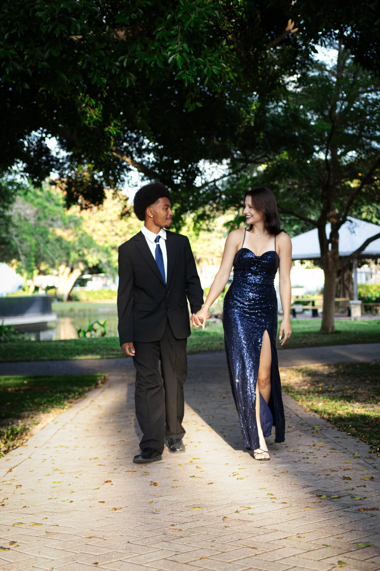 A prom couple walking hand in hand down a tree-lined path lit by warm evening sun
