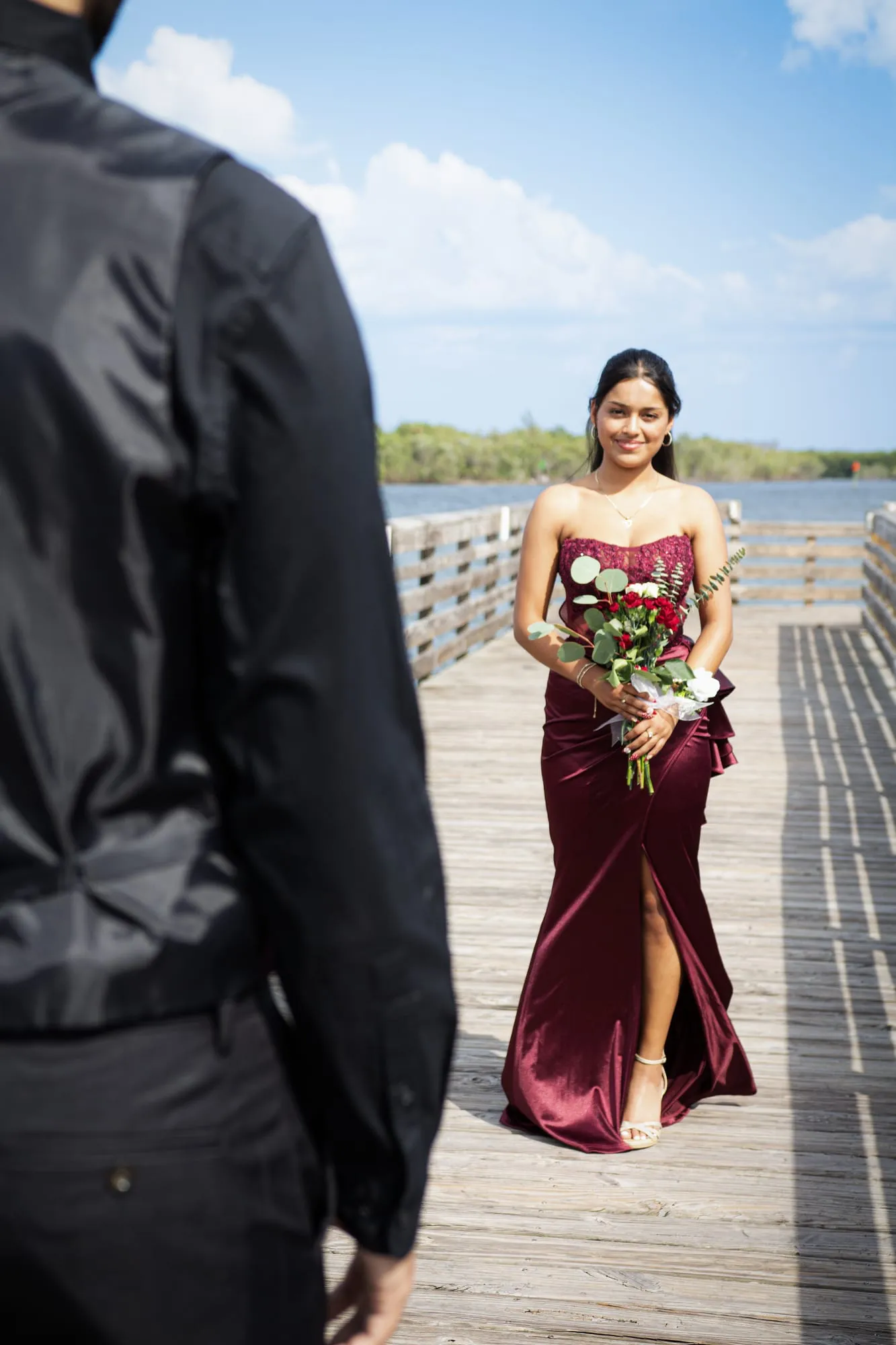 Smiling prom portrait of a girl in a burgundy gown holding a rose bouquet on a sunlit waterfront boardwalk