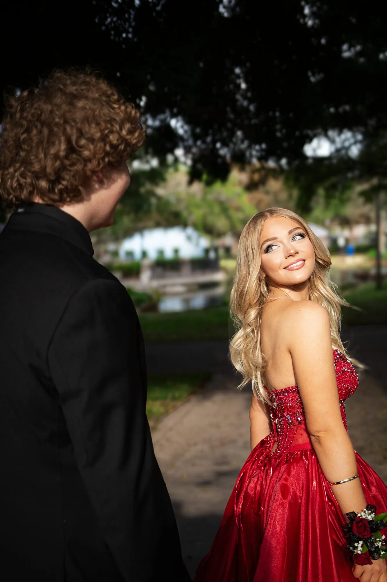 Senior in a dramatic red ballgown beside a moss-draped oak tree, her date in a tuxedo holding her hand
